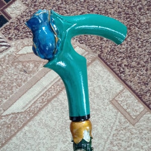 Puede incluir: Un bast&oacute;n de madera con un mango pintado de verde y azul. El mango est&aacute; tallado para parecer una rosa con detalles dorados. El bast&oacute;n est&aacute; decorado con una flor verde y blanca.