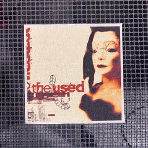 Puede incluir: Parche cuadrado con el texto "the used" en rojo y negro. El parche presenta un retrato estilizado de una mujer con cabello oscuro y una combinación de colores rojo y negro. El fondo es beige neutro.