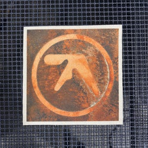 Könnte beinhalten: Quadratischer Aufnäher mit einem kreisförmigen Design in Orange und Braun. Das Design zeigt ein stilisiertes Symbol in einem Kreis, vor einem strukturierten Hintergrund. Der Aufnäher ist von einem weißen Rand umgeben.