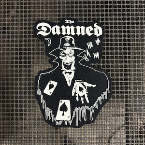 Op de afbeelding: Zwarte patch met een witte illustratie van een sinister figuur met een hoed, die speelkaarten vasthoudt, en de tekst "The Damned". Het ontwerp bevat "HA HA HA" tekstballonnen en druipende details.