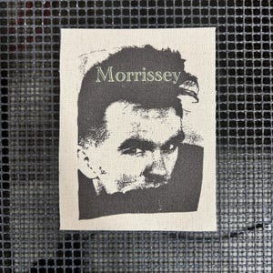 Puede incluir: Un parche rectangular con un retrato en blanco y negro de Morrissey, con el nombre "Morrissey" impreso encima en verde claro. El parche está hecho de una tela de color claro.