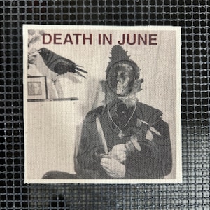 Puede incluir: Un parche cuadrado blanquecino con una imagen en blanco y negro de una persona con vestimenta histórica, sosteniendo un cuchillo. El texto "DEATH IN JUNE" está impreso en rojo oscuro en la parte superior. También se representa un cuervo.