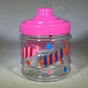 May include: A clear glass jar with a bright pink lid. The jar is decorated with colorful candy-shaped designs in red, pink, orange, and blue, along with polka dots. The jar is empty.