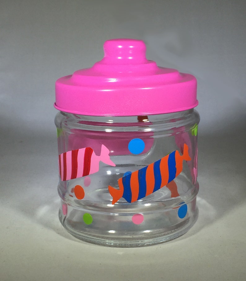May include: A clear glass jar with a bright pink lid. The jar is decorated with colorful candy-shaped designs in red, pink, orange, and blue, along with polka dots. The jar is empty.