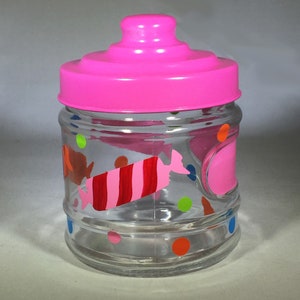 May include: A clear glass jar with a bright pink lid and a pink candy design. The jar is decorated with colorful polka dots and a red and pink candy graphic. The jar is designed to hold small items.