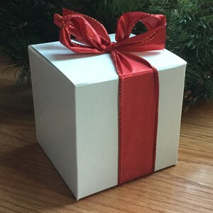 May include: A white square gift box with a vibrant red ribbon and bow. The ribbon wraps around the box, creating a festive look. The background includes greenery and a wooden surface, suggesting a holiday theme.