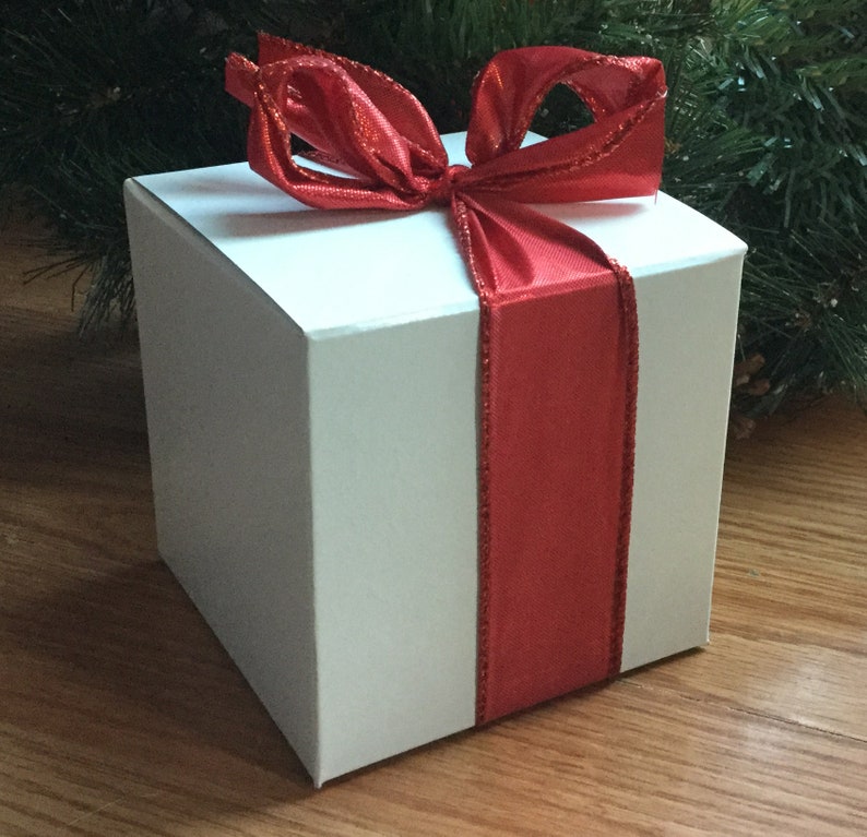 May include: A white square gift box with a vibrant red ribbon and bow. The ribbon wraps around the box, creating a festive look. The background includes greenery and a wooden surface, suggesting a holiday theme.