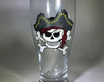 May include: A clear glass beer stein with a hand-painted design of a pirate skull and crossbones. The skull is wearing a black and red pirate hat with yellow trim.