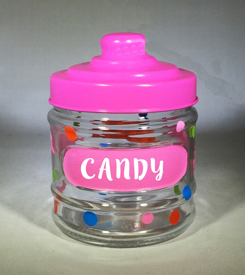 May include: A clear glass candy jar with a bright pink lid and a pink label that reads "CANDY" in white lettering. The jar is decorated with colorful polka dots in blue, green, orange, and pink.