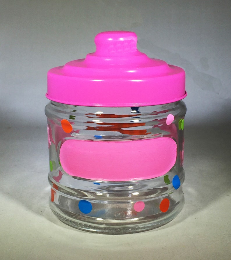 May include: A clear glass jar with a bright pink lid and a matching pink painted label. The jar is decorated with colorful polka dots in orange, blue, green, and pink. The jar is designed to store small items.