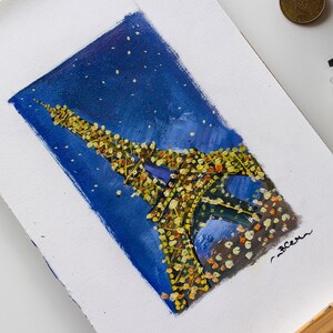 May include: A painting of the Eiffel Tower in Paris, France, with a starry night sky background. The tower is painted in yellow and white, with a blue sky and white stars. The painting is on a white canvas and is on an easel.