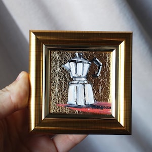 Puede incluir: Una pequeña pintura enmarcada de una cafetera blanca sobre un fondo dorado. La cafetera tiene un asa y un pico negros.