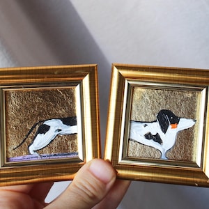 May include: Two small gold framed paintings of black and white dogs on a gold background. The dog in the left frame is running and the dog in the right frame is standing.