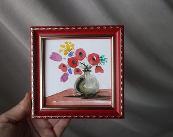 May include: A square framed watercolour painting featuring a bouquet of red, yellow, and purple flowers in a small grey vase. The artwork is set against a reddish-brown surface and is enclosed in a glossy red frame with gold-toned inner trim.
