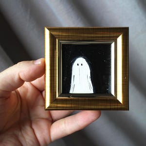 K&ouml;nnte beinhalten: Ein kleines, quadratisches Gem&auml;lde in einem goldenen Rahmen. Das Kunstwerk zeigt eine wei&szlig;e Geisterfigur vor schwarzem Hintergrund. Der Geist hat zwei kleine, schwarze Augen. Der Rahmen hat ein metallisches, strukturiertes Aussehen.
