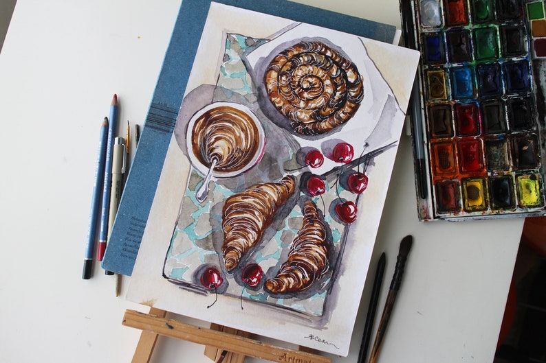 May include: Watercolor painting of a breakfast scene with a cup of coffee, two croissants, and a swirl of pastry. There are also red cherries scattered around the plate. The painting is on a white background with a blue border.