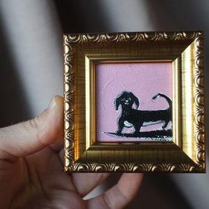 May include: A small framed painting of a black dachshund on a pink background.
