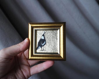 May include: A small, square framed artwork featuring a black and white bird on a gold background. The bird is depicted in a minimalist style. The frame is gold-colored, adding a touch of elegance to the piece. The artwork is held in a hand.
