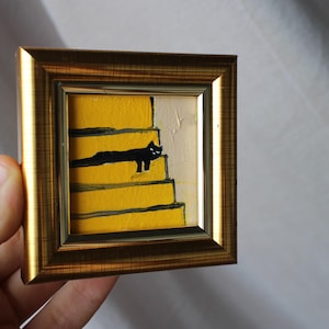 May include: A framed painting of a black cat sitting on yellow stairs. The painting is in a gold frame.