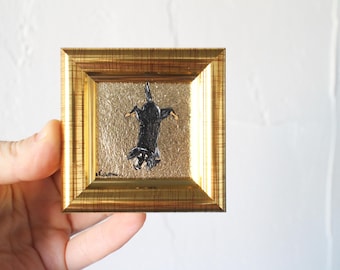 May include: A small framed painting of a black dog on a gold background. The dog is upside down and appears to be looking up.