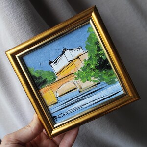 A small, framed painting with a gold-colored frame. The artwork depicts a bridge over water, with buildings and trees. The painting has a blue sky and is held by a hand.