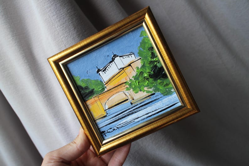 A small, framed painting with a gold-colored frame. The artwork depicts a bridge over water, with buildings and trees. The painting has a blue sky and is held by a hand.