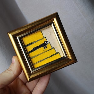 A small framed painting of a black cat walking up yellow stairs. The frame is gold and the background is white.
