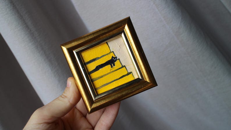 A small framed painting of a black cat walking up yellow stairs. The frame is gold and the background is white.