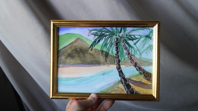 May include: A watercolor painting in a gold frame depicts two palm trees on a beach with a green and brown mountain in the background. The water is a light blue and the sand is a light brown.