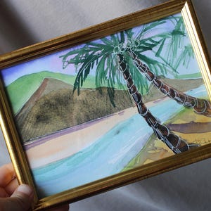 May include: A framed watercolor painting of two palm trees on a beach with blue water and a green and brown mountain in the background.