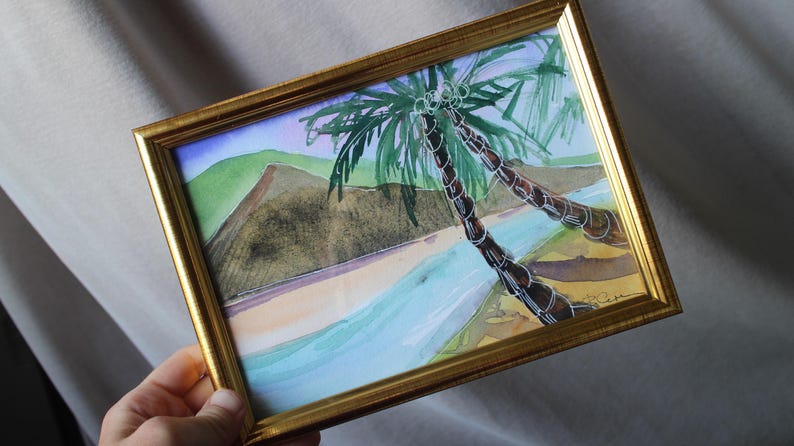 May include: A framed watercolor painting of two palm trees on a beach with blue water and a green and brown mountain in the background.