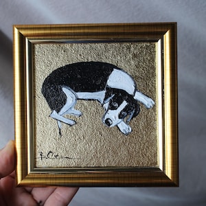 May include: A framed painting of a dog in a playful pose. The dog is black and white, set against a gold background. The frame is gold. The painting is held by a hand.