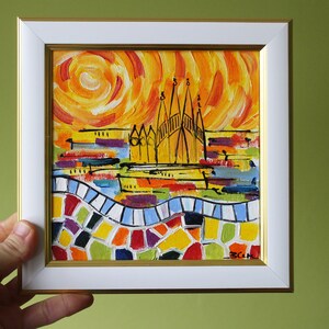 May include: A framed painting depicting a colourful cityscape with a bright orange sun in the background. The cityscape features a tall building with a pointed roof and a mosaic-like pattern at the bottom.