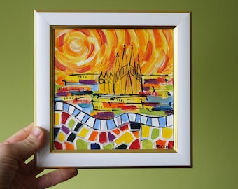 May include: A framed painting of a colorful cityscape with a bright orange sun in the background. The cityscape features a tall building with a pointed roof and a mosaic-like pattern at the bottom.
