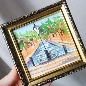 May include: A framed watercolour painting depicting a canal scene with a bicycle parked at the water's edge. The painting includes a lamppost, trees, and buildings in the background.