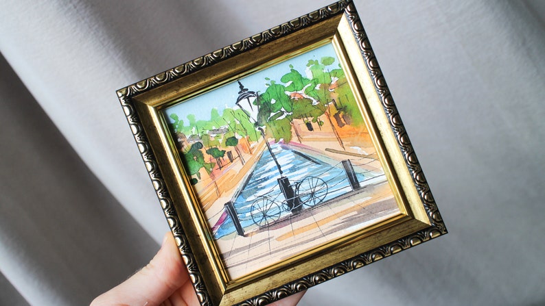 May include: A framed watercolour painting depicting a canal scene with a bicycle parked at the water's edge. The painting includes a lamppost, trees, and buildings in the background.
