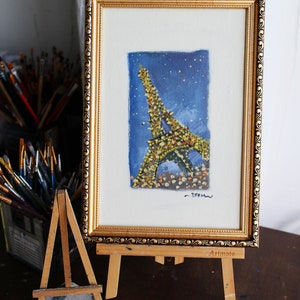May include: A framed painting of the Eiffel Tower in Paris, France. The painting is done in a watercolor style and features a blue background with white stars. The tower is depicted in a yellow and brown color scheme. The painting is signed "Dream" in the bottom right corner and is displayed on a small wooden easel.