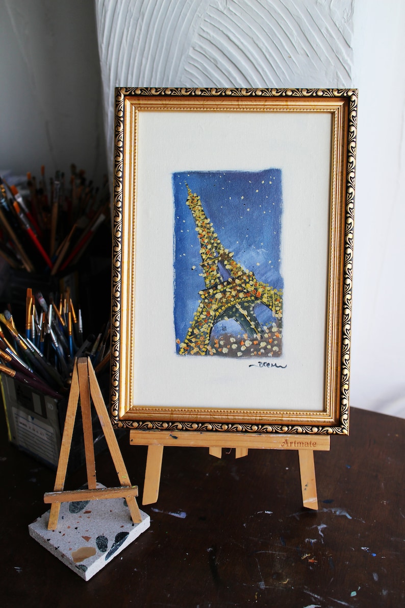 May include: A framed painting of the Eiffel Tower in Paris, France. The painting is done in a watercolor style and features a blue background with white stars. The tower is depicted in a yellow and brown color scheme. The painting is signed "Dream" in the bottom right corner and is displayed on a small wooden easel.