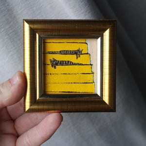 Puede incluir: Una pequeña obra de arte enmarcada con dos gatos estilizados en blanco y negro sobre un fondo amarillo. La obra de arte está en un marco dorado. Los gatos están tumbados en lo que parece ser una escalera.