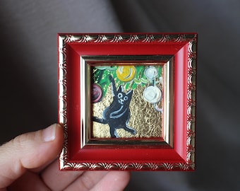 May include: A small square painting in a bright red frame with gold trim. The artwork features a black cat reaching toward a yellow fruit against a textured gold background, with green leaves and white circular elements visible in the composition.