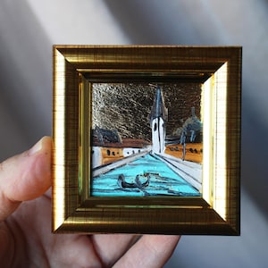 May include: A small framed painting of a blue canal with a white building and a church steeple in the background. The frame is gold and has a textured surface.