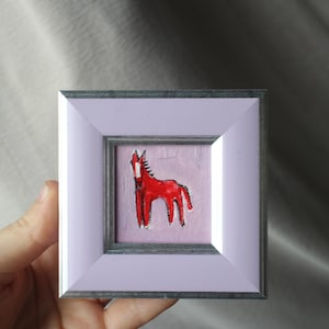 A small, square framed artwork featuring a red horse illustration on a pink background. The frame is a light purple color with a silver inner border. The artwork is held by a hand.
