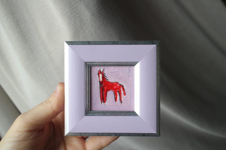 A small, square framed artwork featuring a red horse illustration on a pink background. The frame is a light purple color with a silver inner border. The artwork is held by a hand.