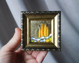 May include: A small framed painting with a gold frame. The painting depicts a landscape with a blue river and yellow trees. The background is gold.