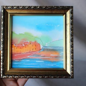 Puede incluir: Una pintura de acuarela de un pequeño pueblo junto al agua, enmarcada en un marco dorado. El pueblo está pintado en tonos de naranja y rojo, con una colina verde en el fondo. El agua está pintada en tonos de azul y verde.