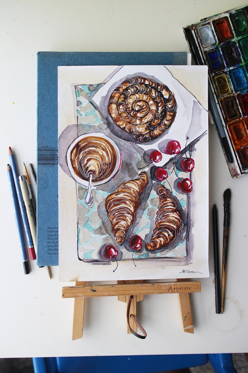 May include: Watercolor painting of a breakfast scene with a croissant, a pastry, a cup of coffee, and cherries on a tiled surface. The painting is on an easel with the word "Artimate" printed on the easel.