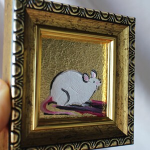 May include: A small framed painting of a white rat with a pink tail sitting on a purple background. The frame is gold with a decorative border.