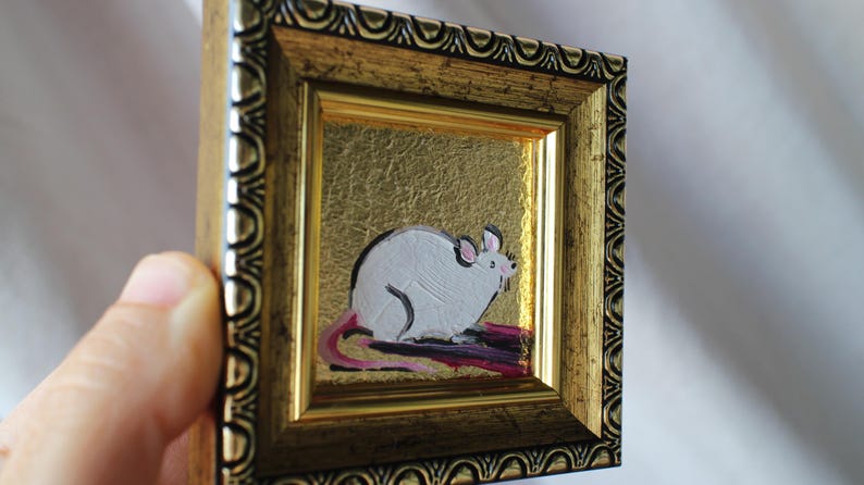 May include: A small framed painting of a white rat with a pink tail sitting on a purple background. The frame is gold with a decorative border.