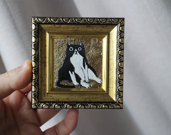 May include: A framed painting of a black and white cat with big eyes sitting on a gold background.