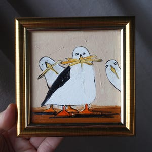 Puede incluir: Pintura enmarcada que representa tres gaviotas blancas estilizadas con detalles negros y patas naranjas. Una gaviota sostiene palos amarillos en el pico. La pintura está enmarcada en un marco dorado.